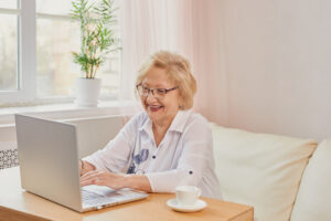 A senior female patient sitting at a desk with cup of coffee looking at her laptop screen cheerfully