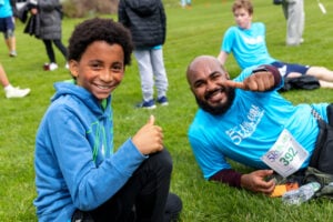 African-American teen and adult in blue shirts sitting on the grass giving a thumbs up